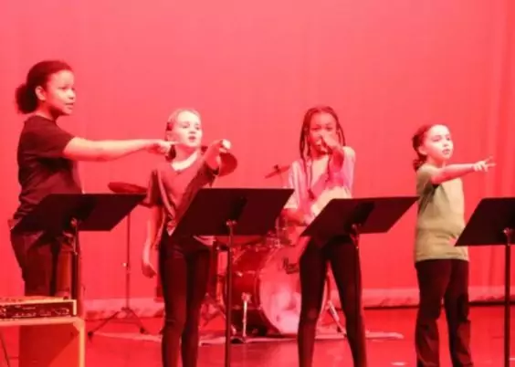 Group of children performing on stage, pointing with enthusiasm under red lighting.