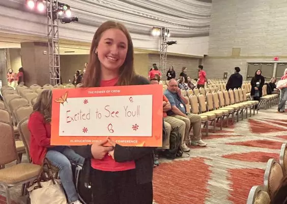 A smiling young woman stands in a large conference hall holding a bright orange sign that reads, “Excited to See You!!” with decorative drawings and text referencing the “EL Education 2023 Conference.” Rows of chairs and event attendees are visible in the background.