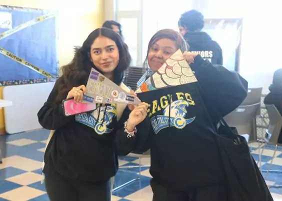 Two students wearing matching “Eagles” hoodies from Capital City Public Charter School smile while holding up their completed creative projects in a classroom setting.