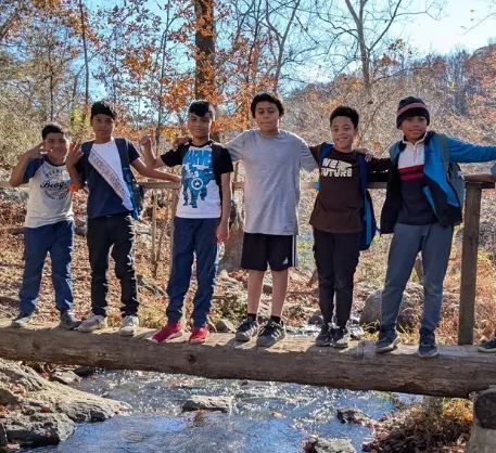 A group of seven boys standing on a log bridge over a creek, smiling and enjoying the outdoors.
