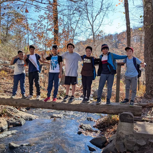A group of seven boys standing on a log bridge over a creek, smiling and enjoying the outdoors.